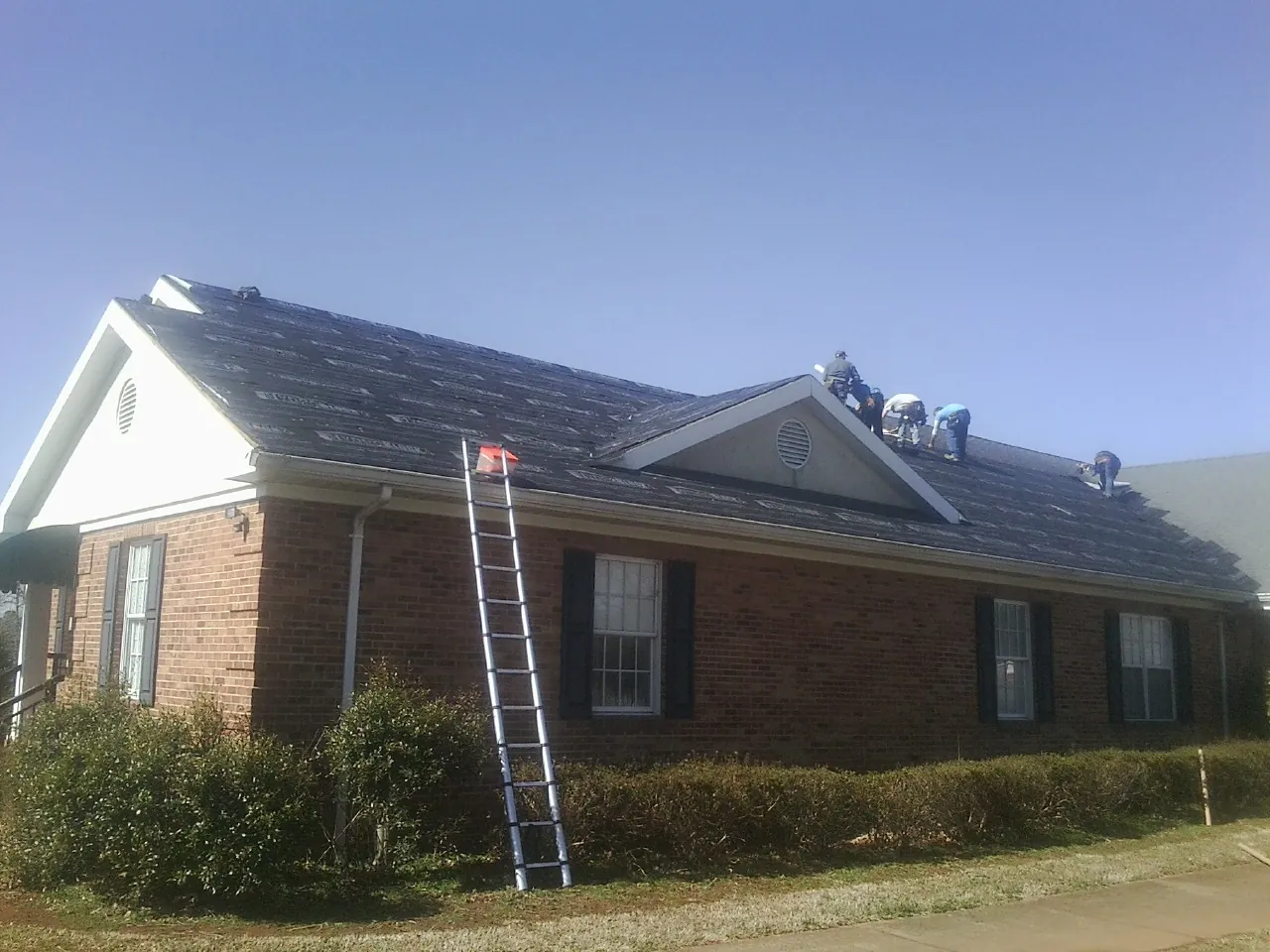 Crew tearing shingles off a brick ranch roof with a ladder set — Metal Roof Repair work by our crew in Shortsville